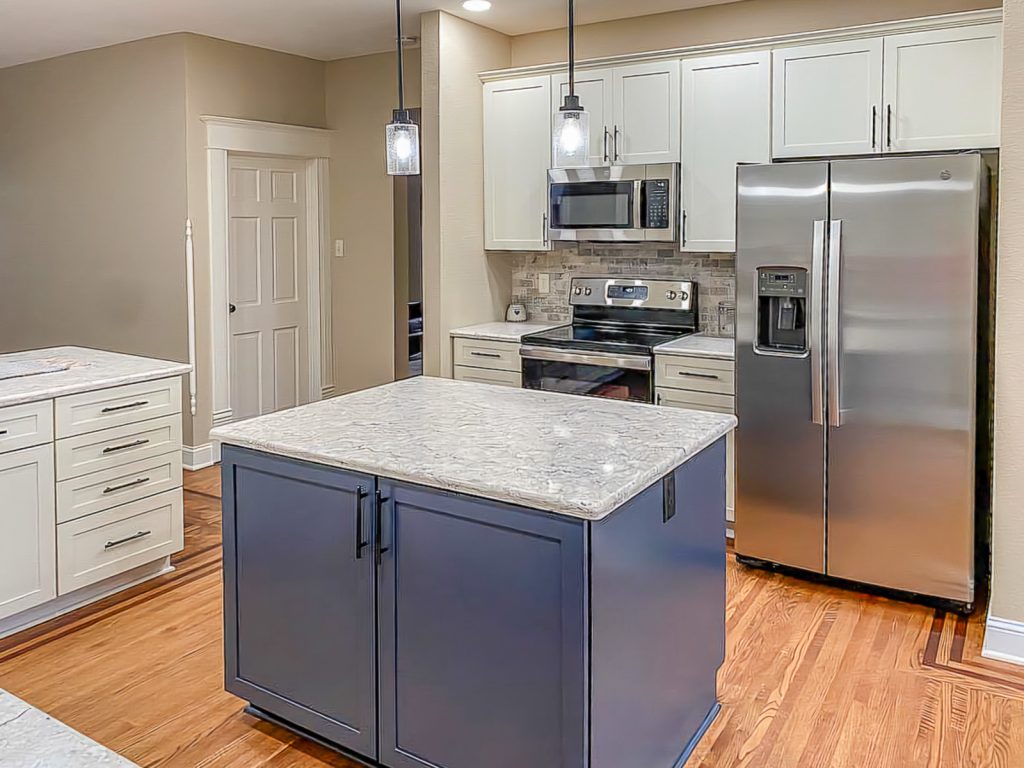Transitional Charm - Elevated Designs: View of blue Island and white cabinet kitchen with marble counters, stainless steel appliances, and wood floors