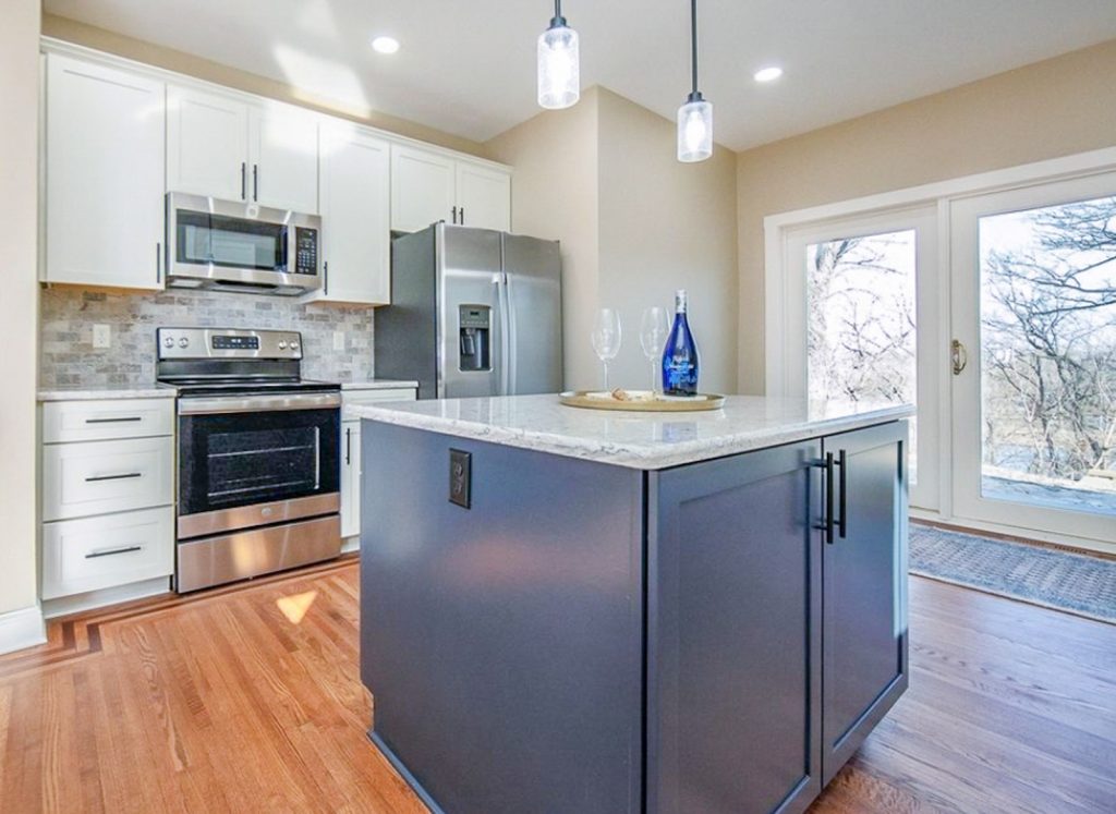 Transitional Charm - Elevated Designs: full View of white cabinet kitchen with marble counters, stainless steel appliances, wood floors. and blue island