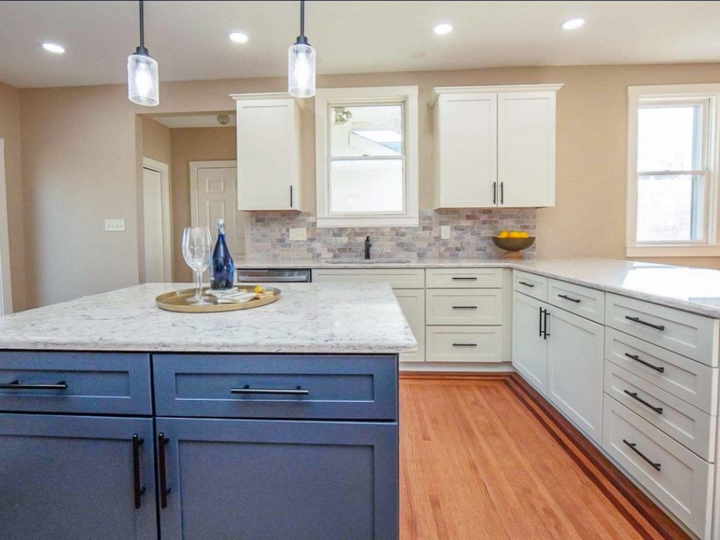Transitional Charm - Elevated Designs: full View of white cabinet kitchen with marble counters, stainless steel appliances, wood floors. and partial blue island