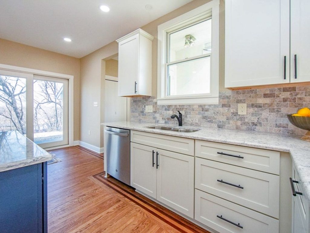 Transitional Charm - Elevated Designs: View of sink area of white cabinet kitchen with marble counters, stainless steel dishwasher
